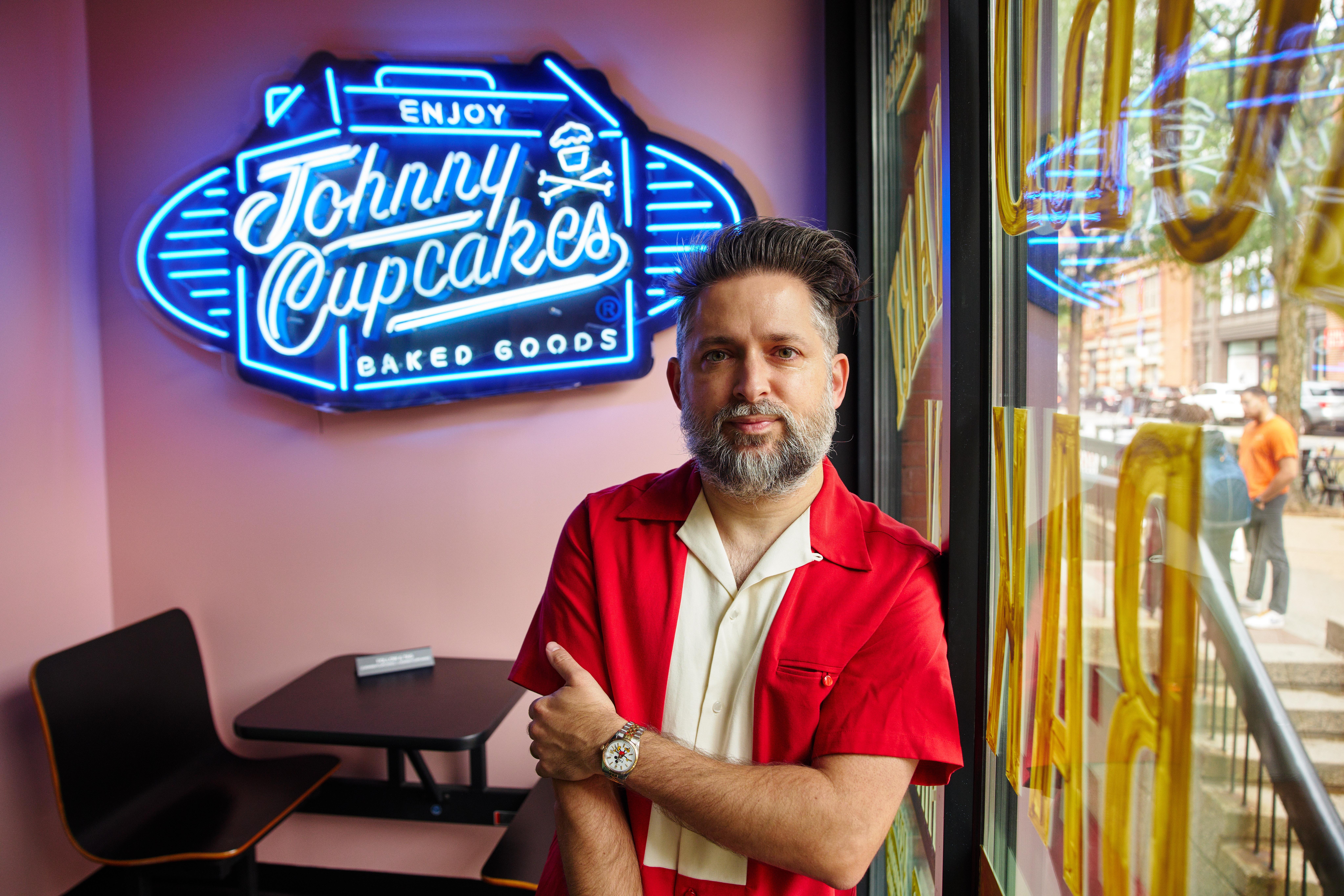 Picture of Johnny Earle with his arm crossed. There is a neon sign in the background that says 'Enjoy Johnny Cupcakes Baked Goods'